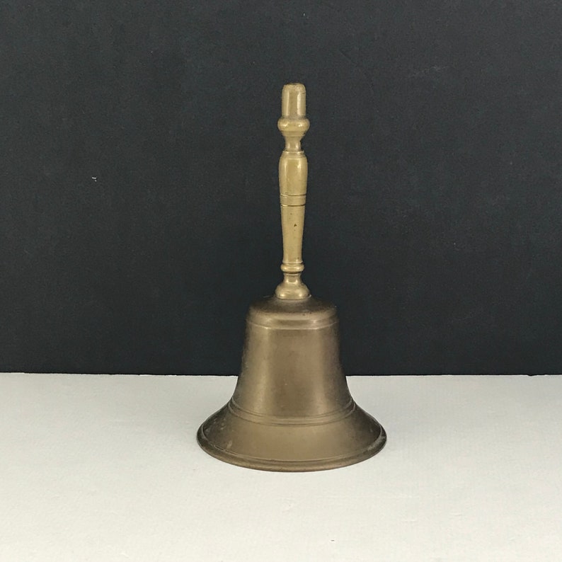 May include: A brass bell with a wooden handle. The bell is a light gold color and has a smooth, polished finish.