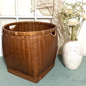 May include: A large, woven brown basket with a square base and two handles. The basket is made of tightly woven bamboo or rattan, with a decorative trim around the top. A white vase with floral patterns and dried flowers is next to the basket.