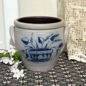 Puede incluir: Una jardinera de gres vintage con un diseño floral azul. La jardinera tiene un borde ancho, dos asas y un interior marrón oscuro. El diseño azul presenta una planta en maceta con hojas y tallos. La jardinera está sobre una superficie estampada.