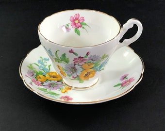 May include: A white teacup and saucer set with a floral pattern. The cup and saucer have a gold trim and feature a variety of colorful flowers, including pink, yellow, and blue.