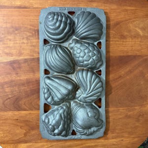 Könnte beinhalten: Eine Backform aus Gusseisen mit Muschelformen. Die Form hat acht Hohlräume, die jeweils zur Herstellung eines anderen Muschelmusters bestimmt sind. Die Form ist dunkelgrau und trägt die Prägung "DED JOHN WRIGHT CO" und "PAT. PEND.".