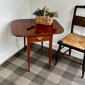 May include: A small, antique mahogany table with a single drawer and brass hardware. A woven basket filled with flowers sits on the table. A black remote control is on the table. A wooden chair with a woven seat is next to the table.