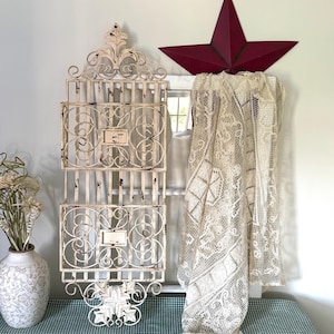 May include: Off-white metal wall organiser with three sections, featuring ornate scrollwork and a distressed finish. A burgundy star and a cream lace curtain are also visible, creating a vintage, farmhouse aesthetic.