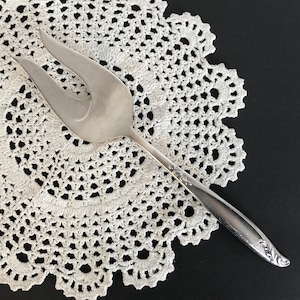 May include: Silver cake server with a decorative handle. The server is on a white crocheted doily.