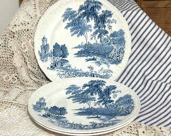 May include: Two white ceramic plates with a blue landscape design. The plates feature a detailed illustration of trees, water, and buildings. The edges of the plates have a scalloped design. The plates are on a white and blue striped background.