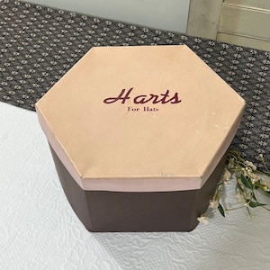 May include: A hexagonal hat box with a brown base and a light pink lid. The lid has the text "Harts For Hats" printed in burgundy.