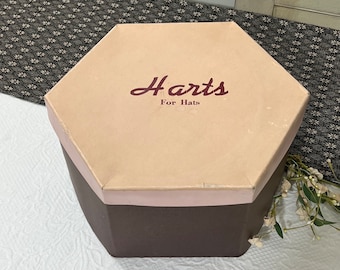 May include: A hexagonal hat box with a brown base and a light pink lid. The lid has the text "Harts For Hats" printed in burgundy.