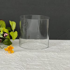 May include: A clear, cylindrical glass vase with an open top and a flat base. The vessel is made of transparent glass and features a simple, smooth design. It is positioned next to a small cluster of artificial green leaves and a yellow flower.