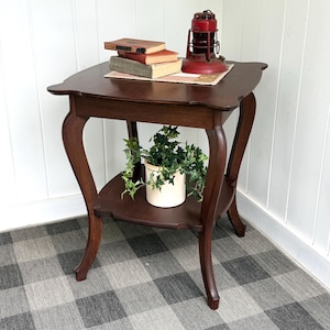 May include: A dark brown, two-tiered side table with curved legs. The top shelf holds a stack of books and a red lantern. The lower shelf displays a potted plant. The table is set on a checkered rug.