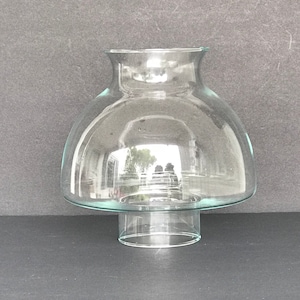 May include: A clear glass lampshade with a wide, rounded base and a cylindrical top. The lampshade is designed to fit over a light bulb.