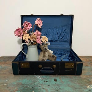 May include: A vintage blue suitcase with a teddy bear and a vase of pink flowers inside. The suitcase has brass hardware and a blue lining.