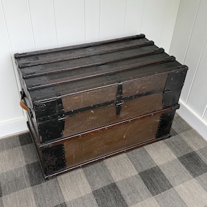 Puede incluir: Un ba&uacute;l de madera vintage con exterior marr&oacute;n oscuro y detalles met&aacute;licos negros. El ba&uacute;l tiene una tapa con bisagras, herrajes met&aacute;licos y un asa de cuero. El ba&uacute;l es rectangular y est&aacute; sobre un suelo a cuadros.