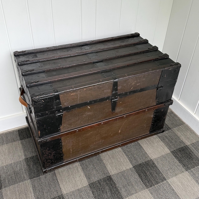 Puede incluir: Un ba&uacute;l de madera vintage con exterior marr&oacute;n oscuro y detalles met&aacute;licos negros. El ba&uacute;l tiene una tapa con bisagras, herrajes met&aacute;licos y un asa de cuero. El ba&uacute;l es rectangular y est&aacute; sobre un suelo a cuadros.