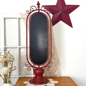 May include: A tall, oval-shaped chalkboard framed in red metal with decorative scrollwork at the top and bottom. The chalkboard is mounted on a sturdy, pedestal-style red base. The frame features a finial at the very top center of the structure.