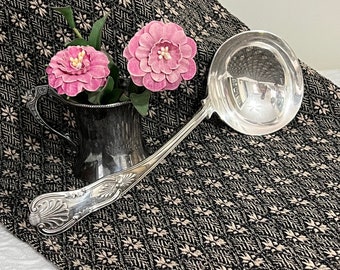 May include: A silver serving ladle with an ornate handle, resting on a black and white patterned fabric. Two pink flowers are in a silver pitcher behind the ladle.
