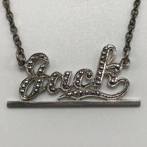 May include: A silver necklace with a pendant that says "Luck" in cursive letters. The letters are made of silver and have small black stones embedded in them. The necklace has a silver chain and a silver bar.