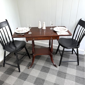 May include: A small, dark wood table with a drawer, set for two with plates, silverware, and glasses. Two black chairs flank the table. The table has a decorative, twisted leg design and a lower shelf. Salt and pepper shakers are also on the table.