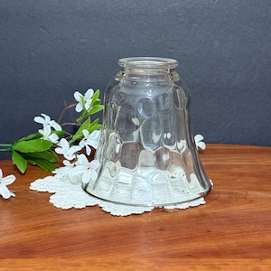 Peut inclure: Un abat-jour en verre transparent avec un motif texturé en nid d'abeille. L'abat-jour a une forme évasée et repose sur un napperon en dentelle blanche. Des fleurs blanches et des feuilles vertes sont visibles en arrière-plan.