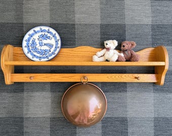 Puede incluir: Una estantería de pared de madera con un plato decorativo azul y blanco, dos osos de peluche y un cuenco de cobre. La estantería tiene una parte superior festoneada y dos ganchos debajo. El plato presenta un diseño de pájaro.