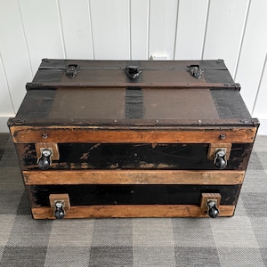 Puede incluir: Un ba&uacute;l antiguo con tapa marr&oacute;n oscuro y base negra. El ba&uacute;l tiene herrajes met&aacute;licos, detalles de madera y ruedas peque&ntilde;as. El ba&uacute;l es rectangular y parece estar hecho de madera y metal.