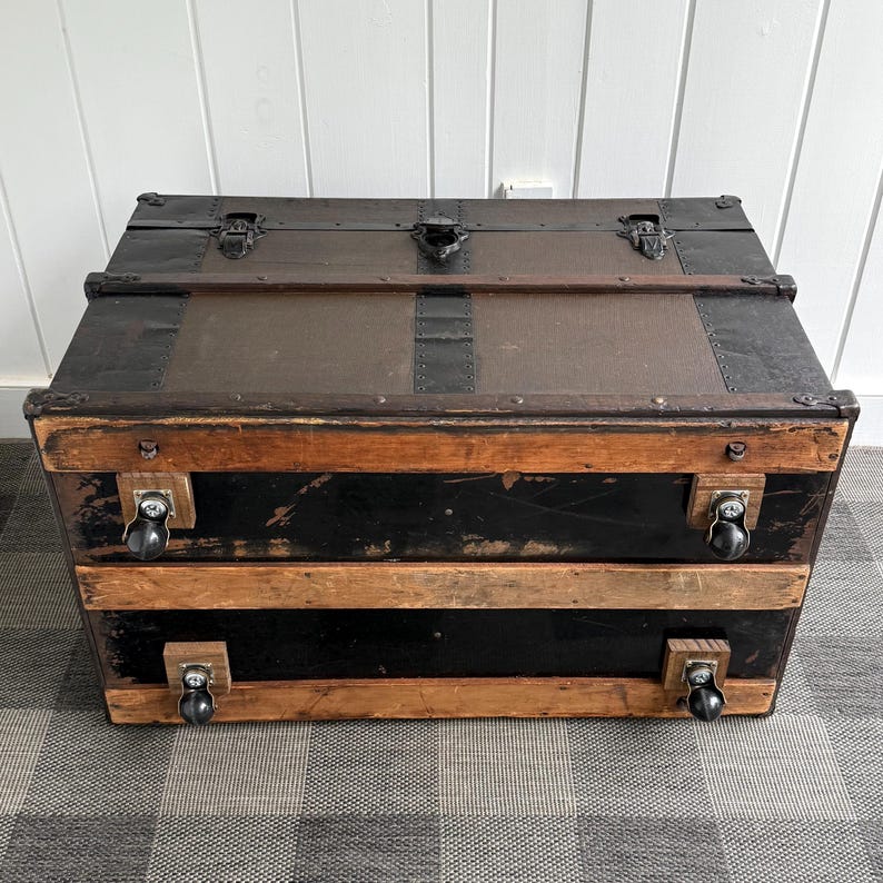 Puede incluir: Un ba&uacute;l antiguo con tapa marr&oacute;n oscuro y base negra. El ba&uacute;l tiene herrajes met&aacute;licos, detalles de madera y ruedas peque&ntilde;as. El ba&uacute;l es rectangular y parece estar hecho de madera y metal.