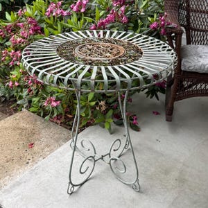 May include: A round, vintage metal patio table with a distressed, light green finish. The tabletop features a decorative, openwork design with a central, ornate medallion. The table has three curved legs and is surrounded by pink flowers.