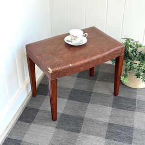 May include: A vintage brown leather-topped stool with four wooden legs. A floral teacup and saucer sit on the stool's surface. The stool is placed on a checkered gray and white rug.
