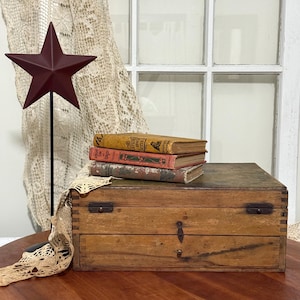 May include: A burgundy metal star on a black pole stands next to a wooden box with three antique books. The books are stacked on top of the box, which has metal hardware and a keyhole. A cream-colored lace curtain hangs in the background.