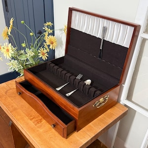 May include: A wooden flatware chest with a hinged lid and a pull-out drawer. The interior is lined with black fabric featuring slots for silverware, including a fork and spoon. The lid interior has white pleated fabric and a single utensil holder.