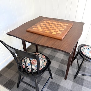 May include: A square wooden game table with a dark brown finish and a built-in checkerboard top featuring alternating light and dark wood squares. Two black wooden chairs with round, floral-patterned needlepoint seat cushions are positioned nearby.