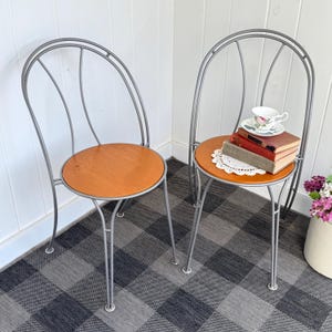May include: Two vintage metal chairs with wooden seats. The chairs have a silver finish and a curved back design. One chair holds a stack of books, a doily, and a teacup and saucer. The seats are a warm, natural wood tone.