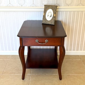 May include: A dark brown wooden end table with a single drawer featuring a brass-toned handle and a lower shelf. An oval-shaped picture frame with an ornate metallic border sits on the flat top surface of the table.