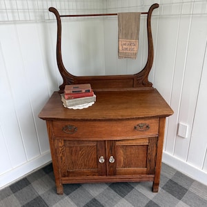 May include: A wooden washstand featuring a curved towel rack, a single drawer with two metal pulls, and a lower cabinet with two doors and clear glass knobs. A brown and white striped towel with the text Live a little Love a lot hangs above.
