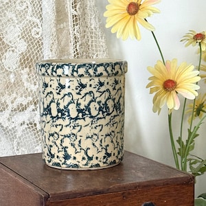 May include: A cylindrical stoneware crock with a cream-colored base and a dark blue, abstract pattern. The crock sits on a wooden surface, with yellow flowers in the background. The crock has a rolled rim.