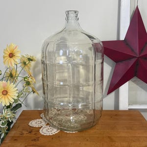 Könnte beinhalten: Eine große, klare Glasballonflasche mit einem gitterartigen Prägemuster auf dem zylindrischen Körper, einem schmalen Hals und einer kleinen kreisförmigen Öffnung an der Oberseite. Das Glas ist transparent und weist vertikale und horizontale Linien auf.