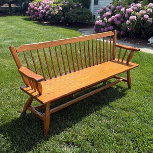 May include: A wooden bench with a slatted back and armrests. The bench is made of light brown wood and is set on a grassy lawn. The bench has a classic design, with a curved back and angled legs.