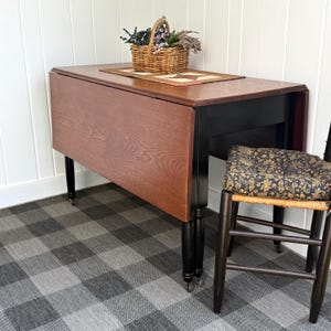 May include: A vintage drop-leaf table with a reddish-brown tabletop and black legs. A woven basket with dried flowers sits on top. The table is paired with a wooden chair with a patterned cushion. The table measures approximately 48 inches long when fully extended.