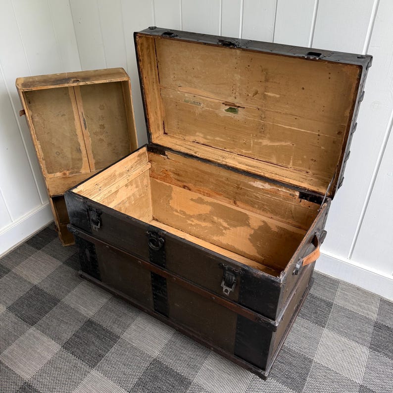 Puede incluir: Un ba&uacute;l de madera antiguo con la tapa abierta, revelando un interior espacioso. El ba&uacute;l es de color marr&oacute;n oscuro con detalles met&aacute;licos negros y asas de cuero. Una caja de madera m&aacute;s peque&ntilde;a est&aacute; cerca. El ba&uacute;l est&aacute; sobre un suelo a cuadros.