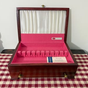 May include: A dark wood cutlery chest with a hinged lid, featuring a white satin-lined interior and a bright pink fabric base. The interior includes a slotted rack for utensils and a small white card with the text 1847 and a guarantee.