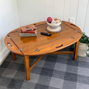 May include: An oval wooden coffee table with a removable tray top featuring brass hinges and handles. The table surface holds two stacked books, a black remote control, and a white ceramic bowl filled with decorative spheres on a round mat.