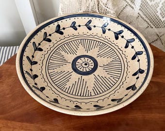 Könnte beinhalten: Eine beige Keramikschale mit blauem Blumendesign. Die Schale hat ein zentrales kreisförmiges Muster mit radialen Linien und einen Rand aus stilisierten Blättern. Der Rand ist ebenfalls mit blauen Blättern und Punkten verziert.