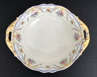 May include: A white porcelain serving bowl with gold and blue floral accents and gold handles. The bowl has a scalloped edge and a floral pattern around the rim.