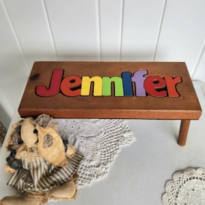 Puede incluir: Un taburete de madera con el nombre "Jennifer" escrito en piezas de rompecabezas de colores. El taburete tiene un acabado de madera natural y dos patas.