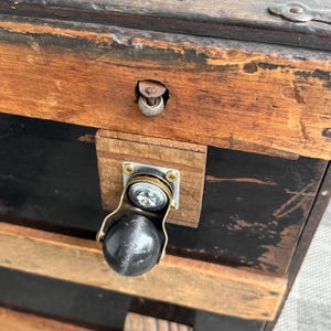 Puede incluir: Primer plano de un ba&uacute;l de madera vintage con acabado negro y marr&oacute;n. El ba&uacute;l presenta una rueda met&aacute;lica con una bola negra y un pestillo met&aacute;lico. La madera tiene vetas visibles y signos de desgaste.
