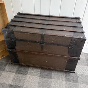 Puede incluir: Un ba&uacute;l de madera vintage de color marr&oacute;n oscuro, reforzado con bandas y protectores de esquina de metal negro. El ba&uacute;l tiene una tapa con bisagras y cierres de metal, ideal para el almacenamiento. El ba&uacute;l est&aacute; colocado sobre una superficie a cuadros.