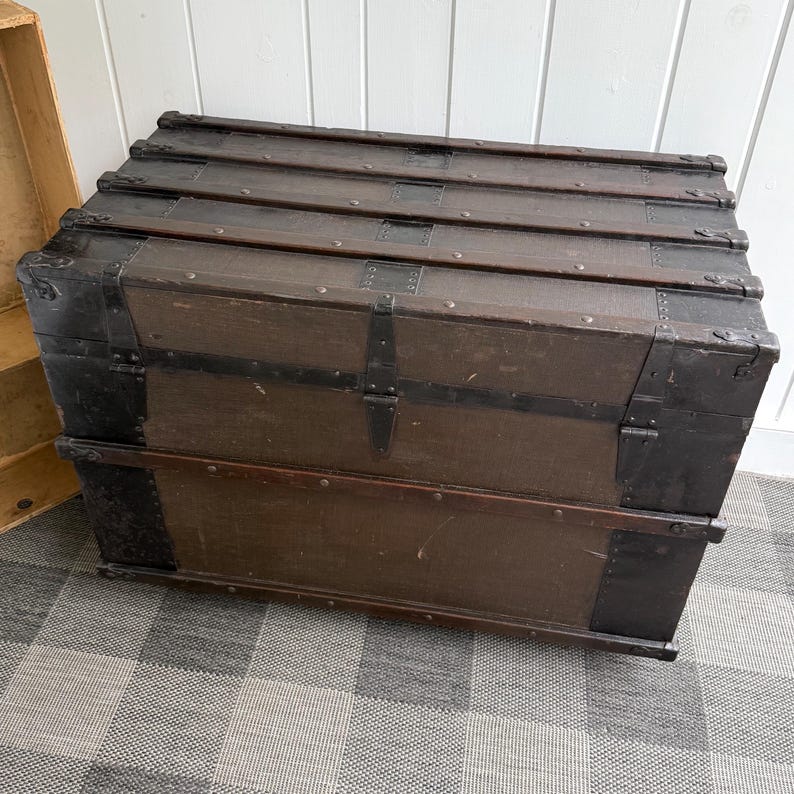 Puede incluir: Un ba&uacute;l de madera vintage de color marr&oacute;n oscuro, reforzado con bandas y protectores de esquina de metal negro. El ba&uacute;l tiene una tapa con bisagras y cierres de metal, ideal para el almacenamiento. El ba&uacute;l est&aacute; colocado sobre una superficie a cuadros.