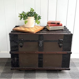 Puede incluir: Un ba&uacute;l antiguo con paneles de madera marr&oacute;n oscuro y detalles de metal negro. Un tablero de ajedrez, una planta en maceta y varios libros descansan encima. El ba&uacute;l tiene cierres y protectores de esquina de metal.