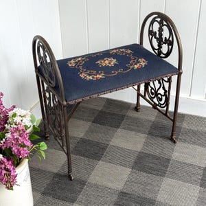 May include: An antique bench with a dark blue upholstered seat featuring a floral design. The bench has a wrought iron frame with decorative scrollwork on the back and sides. The bench is a dark bronze color.