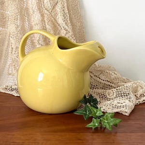 May include: A yellow ceramic pitcher with a rounded body, a curved handle, and a spout. The pitcher is sitting on a wooden surface with green leaves and a beige lace cloth in the background.
