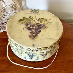 May include: Round decorative box with a lid featuring a watercolor illustration of purple grapes and green leaves. The box is labeled "Chateau Montagne Cabernet Sauvignon" and has a white rope handle.